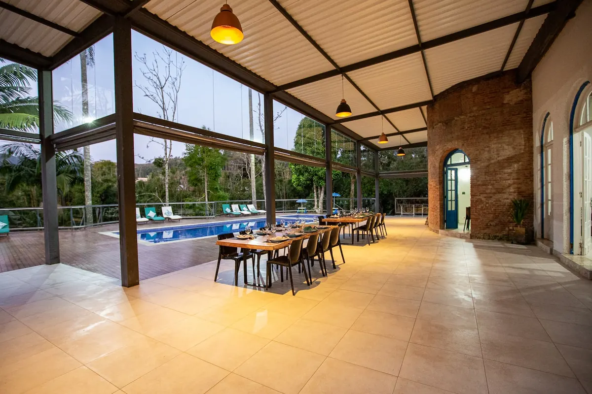 Salão coberto de vidro e aço, aberto para a piscina e a mata